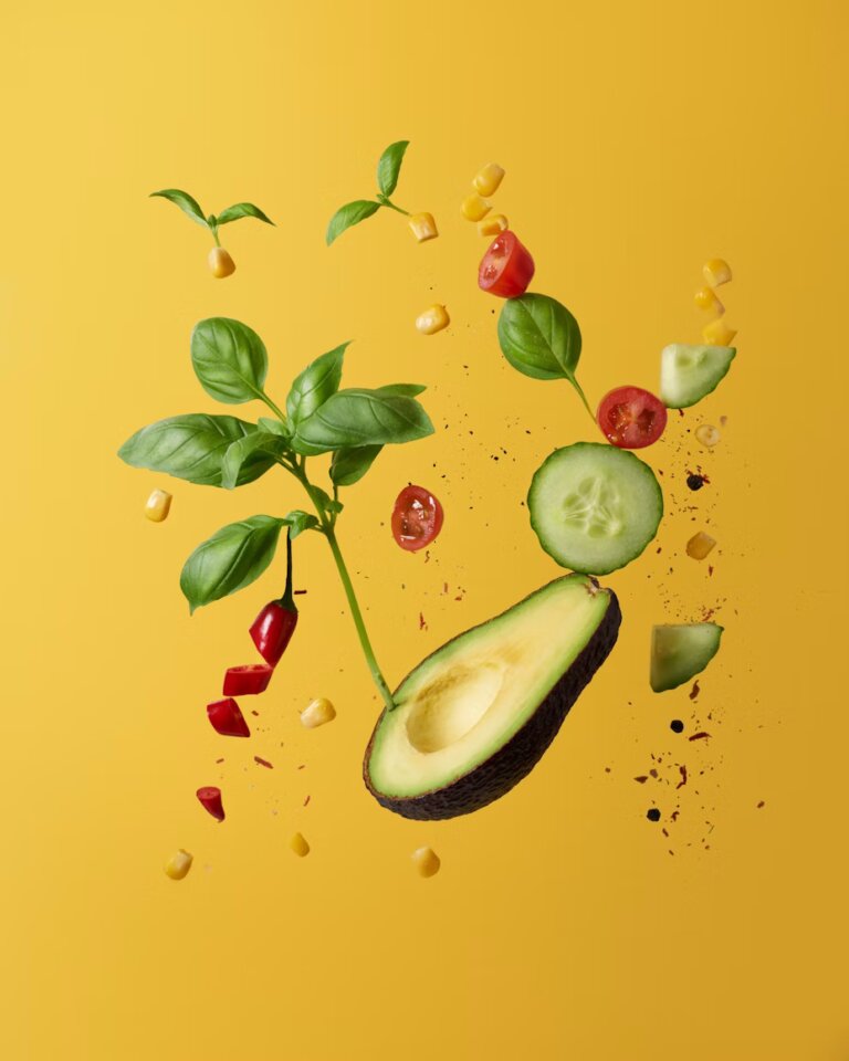 Halbierte Avocado, Gurkenscheibe, Basilikumblätter, Kirschtomatenhälften, Maiskörner und rote Paprikastücke schweben vor gelbem Hintergrund.
