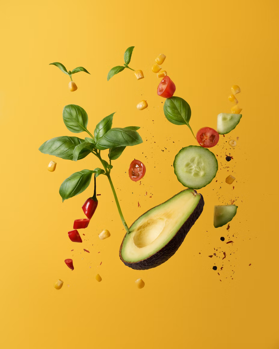 Halbierte Avocado, Gurkenscheibe, Basilikumblätter, Kirschtomatenhälften, Maiskörner und rote Paprikastücke schweben vor gelbem Hintergrund.