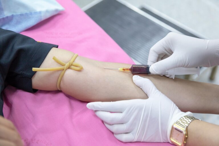 Ein medizinischer Mitarbeiter mit Handschuhen entnimmt einer Person mit einer Spritze Blut aus dem Arm; oberhalb der Einstichstelle wird ein Stauschlauch angelegt.
