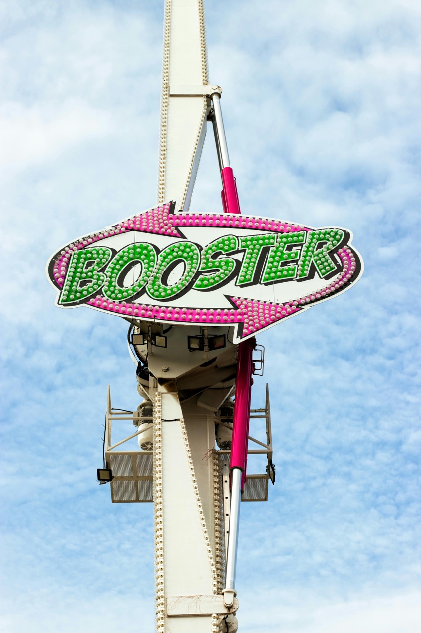 Leuchtschild mit dem Schriftzug 'BOOSTER' in grüner Schrift auf pink-weißem Pfeil vor blauem Himmel